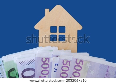 model of a toy wooden house on a blue background. Under it are euro and dollar bills. Close-up, horizontal photo. Idea - buying, renting, selling real estate.