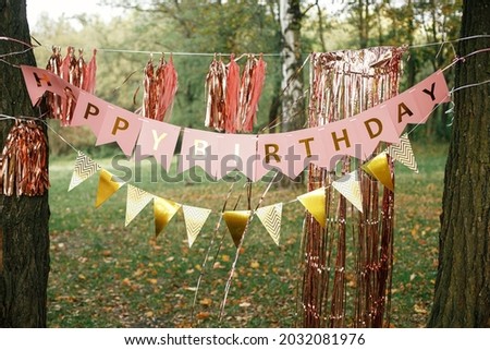 Stylish happy birthday garland hanging in park. Modern rose gold decor, tassel garland and happy birthday banner hanging on trees picnic party. Birthday celebration outdoor