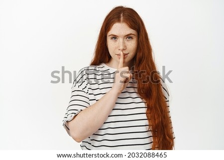 Hush, dont tell anyone. Mysterious redhead girl shushing, make shh sign with finger on lips and smiling, telling secret, asking keep quiet, standing against white background