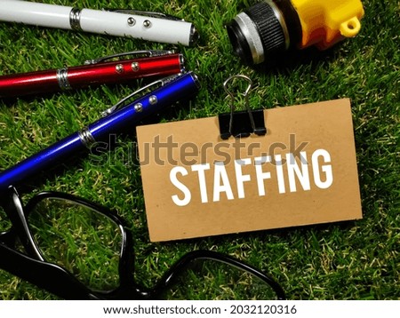 Business concept.Text STAFFING writing on brown card with small camera,glasses and pen on grass background