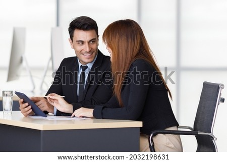 Caucasian happy handsome successful businessman in black formal suit smiling listening to Asian pretty businesswoman who use pencil pointing at tablet screen explaining important business information.