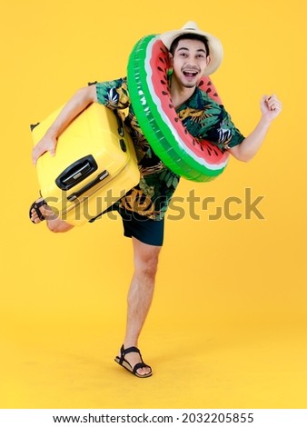 Joyful young Asian man in colorful Hawaiian shirt stands on one leg holds yellow suitcase and swim ring. Full body funny studio portrait on yellow background. Summer holiday travel concept