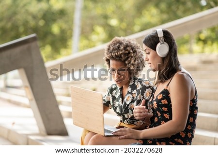 multiracial female friends using laptop smiling looking at screen, wearing headphones and casual clothes, outdoors