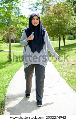 Picture of a young Muslim woman outside a green nature park doing exercises with a skipping rope.
