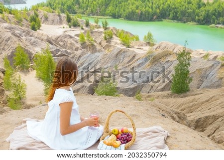 teenage girl in white dress is sitting on edge of cliff next to picnic basket, background of stunningly beautiful landscape with rocks and lake outdoor