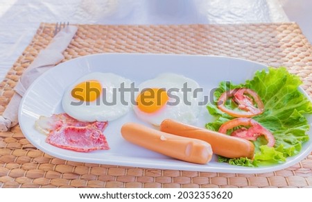 Pictured above of breakfast in white on a dining table with two fried eggs and two hot dogs and bacon and vegetables on the side.