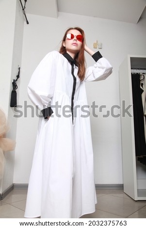 Glamorous model wearing white dress and red eyeglasses, posing over white wall background indoor. Female high fashion accessory concept. Fashion shooting in studio. Professional model, copy space
