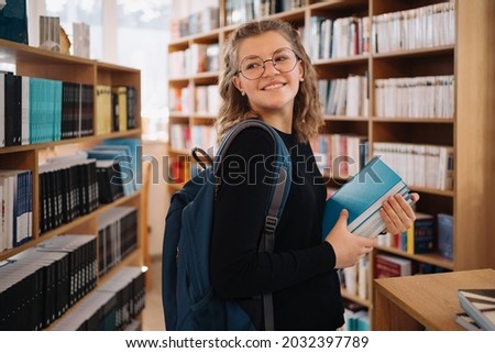 Happy teenage girl or student wearing glasses taking book from shelf in library, wear backpack- People, knowledge, education and school concept