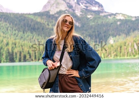 Outdoor picture of pong blonde tours woman having vacation at Montenegro Black lake, posing near forest and mountains, casual outfit.