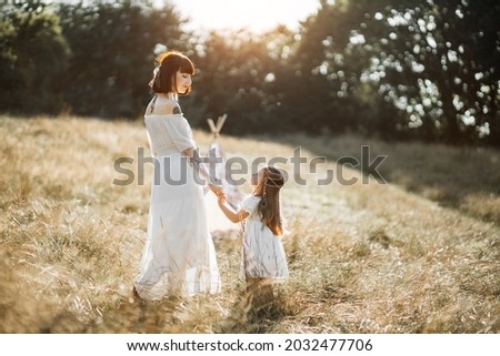 Happy motherhood, summertime. Caring gorgeous mother walking with her cute child girl at sunset in wild field. Indian American mom and daughter wearing boho white dresses and feather hair accessories