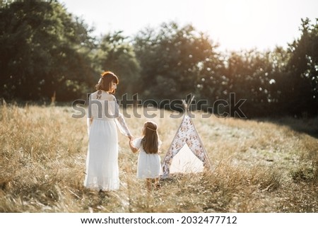 Back rear view of stunning mother and little daughter dressed in native american style, dress and hair feathers, holding hands and looking each other, while walking at summer field