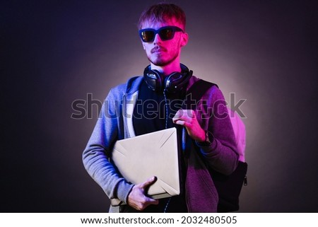 Neon portrait of a bearded man wearing headphones on the neck, parka, backpack, mate sunglasses holding a gamer laptop in hands. Hacker portrait
