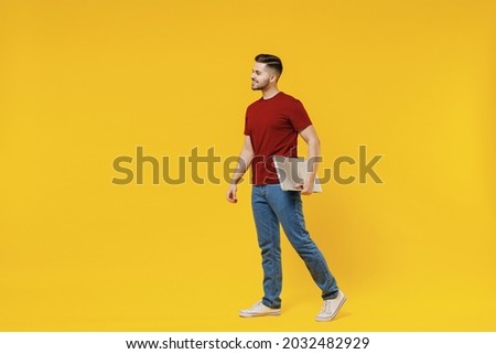 Full length smiling happy young man in red t-shirt casual clothes hold closed laptop pc computer work look aside isolated on plain yellow color wall background studio portrait People lifestyle concept