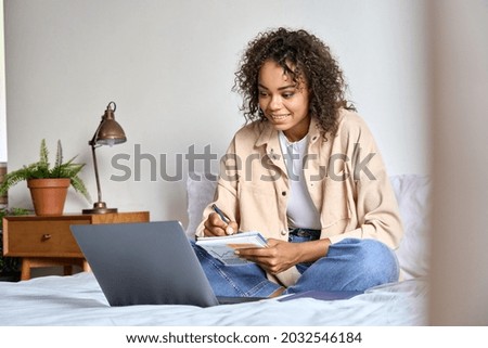 Young happy smiling Afro American mixed race female student watching online class on laptop computer writing notes sitting on bed at modern bedroom apartment. Remote distant e learning work concept.