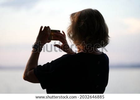 Silhouette of elderly woman with mobile phone taking photo of beautiful sunset at lake Bodensee on sunny summer evening. Photo taken August 15th, 2021, Bregenz, Austria.