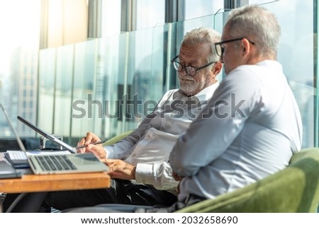 Picture of young business man taking to his older business partner. They are in white shirt and black tie. They are in a hotel lobby. 