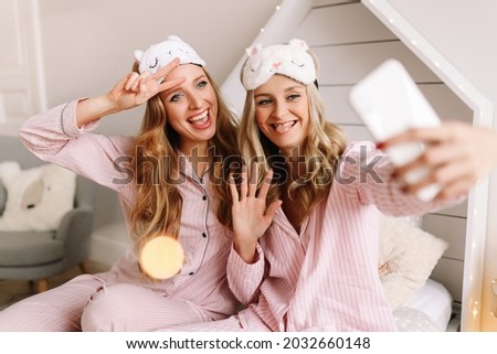 Two happy women friends in pajamas and sleep masks have fun smile take selfies communicate via video communication using a mobile phone sitting on the bed at home during the holidays, selective focus