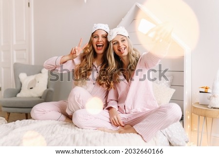 Two happy women friends in pajamas and sleep masks have fun smile take selfies communicate via video communication using a mobile phone sitting on the bed at home during the holidays, selective focus