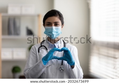 Happy young indian ethnicity female gp doctor infectious disease specialist in disposable gloves and protective facial mask making heart sign, expressing support to ill patient, charity concept.