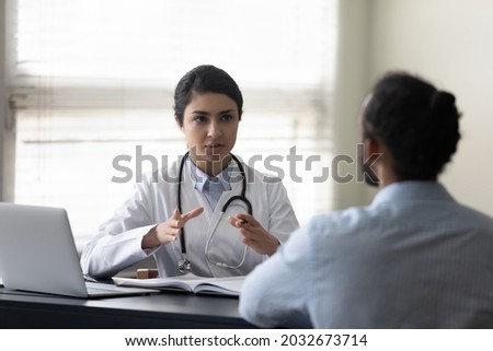 Concentrated young female indian doctor physician giving healthcare consultation to african american male patient, explaining medical insurance benefits or illness treatment at checkup meeting. Royalty-Free Stock Photo #2032673714
