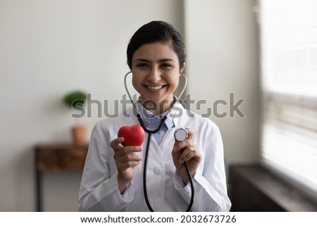Smiling young professional female indian ethnicity cardiologist wearing stethoscope, showing heart figure to camera, reminding regular examination checkup meeting, cardiovascular disease prevention. Royalty-Free Stock Photo #2032673726