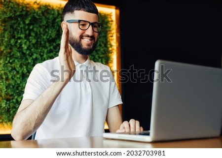 Portrait of cheerful young man in white t-shirt and business glasses using laptop and having video call on laptop computer in cafe, concept of work, learning, remote work Royalty-Free Stock Photo #2032703981