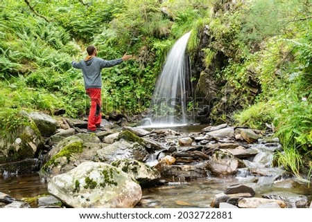 Back view man with raised hands enjoying a waterfall surrounded of vegetation. 