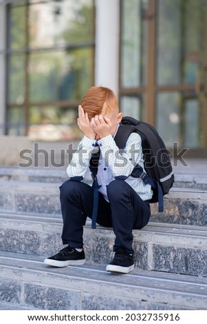 A schoolboy with red hair sits on the stairs by the school, burying his face with his hands and crying Royalty-Free Stock Photo #2032735916