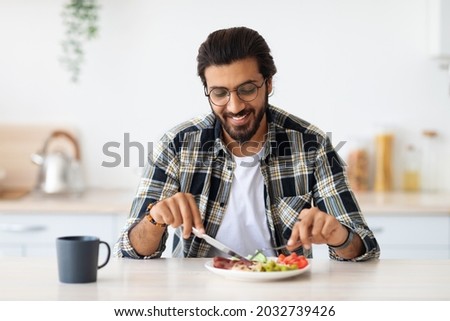 Positive arab guy hipster having healthy breakfast at home, eating fresh vegetables, sausages, drinking coffee, sitting at kitchen table, copy space. Handsome indian man starting day with nice meal Royalty-Free Stock Photo #2032739426