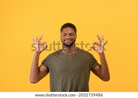 Stress relief concept. Young black man meditating with closed eyes, keeping calm on yellow studio background. African american guy practicing yoga, searching for inner balance