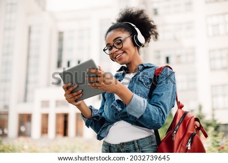 E-Learning. Happy Black Female Student Using Tablet Computer Wearing Headphones Standing Near Modern University Building Outside. Teenager Girl Browsing Internet Outdoors. Modern Online Education Royalty-Free Stock Photo #2032739645