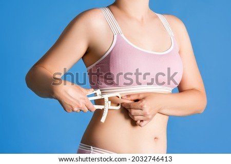 Slim woman in pink underwear measuring her body fat with caliper on blue background, body care concept