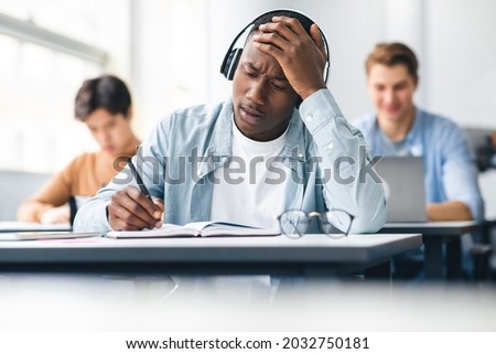Portrait of shocked black student in headphones sitting at table in classroom, touching forehead in panic, writing entrance exam looking at hard questions or test result, thinking about right answer Royalty-Free Stock Photo #2032750181