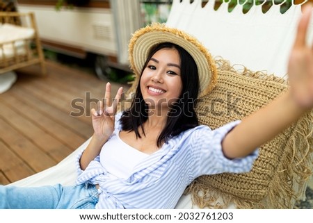 Happy young Asian woman taking selfie while lying in hammock near RV on autumn vacation in countryside. Cheerful millennial lady making photo of herself, showing peace gesture, relaxing outdoors