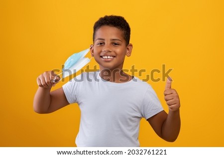 COVID-19 lockdown, pandemic restrictions for kids, children, teens cocnept. Cheerful preteen black boy taking off his protective face mask, showing thumb up, yellow studio background, copy space