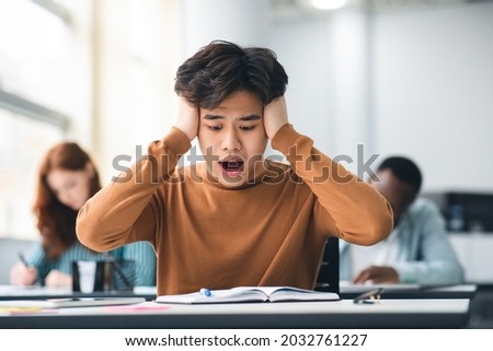 Portrait of shocked asian freshman sitting at table in classroom, grabbing clasping his head in horror and panic, looking at hard exam questions or test result, unhappy about deadline or low score Royalty-Free Stock Photo #2032761227