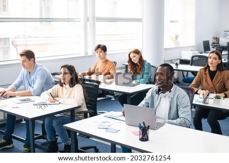 Lecture Concept. Group of interested multicultural mixed race group of students sitting at tables with pc in modern classroom, listening to teacher, taking notes writing in notebooks, back to study