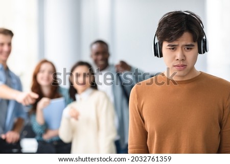 Group Conflict. Sad asian guy in headphones standing away alone, suffering humiliation and public disgrace, crowd of bad people bullying pointing fingers at victim, laughing. Racial Discrimination Royalty-Free Stock Photo #2032761359