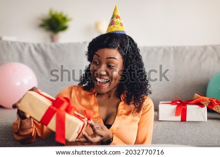Excited Black Woman Opening Wrapped Birthday Gift Box Celebrating Her B-Day And Receiving Presents At Home. Delivery Service, Holiday Celebration Concept. Front View