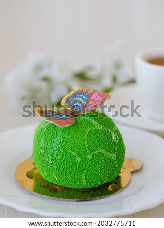 Tender green mousse cake decorated with blue pink edible wafer paper butterfly on plate, cup of tea and white flowers on white background. Selective focus, vertical