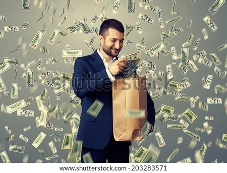 smiley glad businessman standing under money rain and putting dollar's in the paper bag