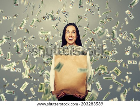 excited laughing businesswoman standing under dollar's rain and holding big paper bag with money