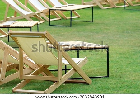 Many empty white deck chairs with tables in lawn is surrounded by shady green grass. Comfortable on outdoor patio chairs in garden.  Lawn chairs in the park. Sunbeds in the garden. Selective focus.