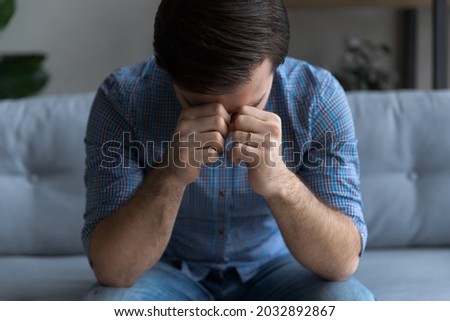 Sad frustrated millennial man suffering from stress, depression at home, sitting on couch, touching head, feeling headache, migraine, coping with loss, grief, trauma. Nervous breakdown, crisis concept Royalty-Free Stock Photo #2032892867