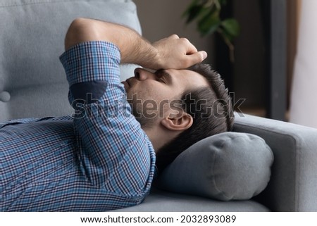 Tired frustrated millennial man lying on sofa at home, thinking over problems, feeling stress, fatigue, touching head, suffering from headache, coping with trauma, loss, addiction, depression, apathy Royalty-Free Stock Photo #2032893089