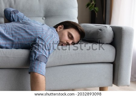 Tired millennial guy in casual sleeping at daytime due to overwork, fatigue, stress, sleepless night. Lazy sleepy bored guy lying on couch with closed eyes, relaxing at home. Energy recreation concept Royalty-Free Stock Photo #2032893098