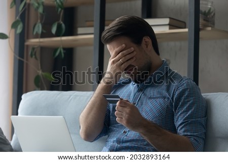 Upset desperate man having problems with wasting money, overspending,, scam and digital fraud. Frustrated disappointed shopaholic, bankrupt holding credit card at laptop, touching face Royalty-Free Stock Photo #2032893164