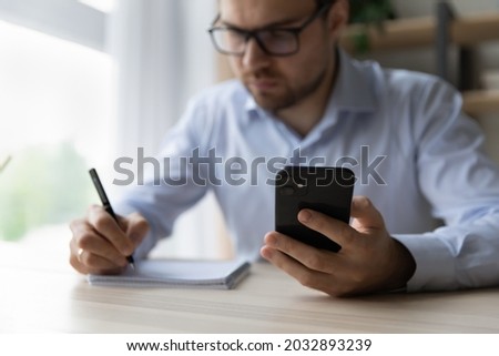 Focused serious millennial business man using mobile phone and taking notes. Male adult student watching learning webinar, online workshop, virtual training on smartphone, writing summary. Close up Royalty-Free Stock Photo #2032893239