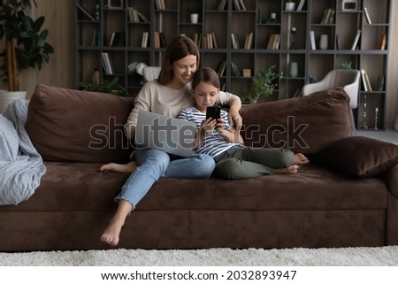 Addicted to modern technology happy young mother and adorable little kid daughter using different gadgets, playing entertaining spending time online resting on comfortable sofa in living room. Royalty-Free Stock Photo #2032893947