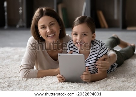 Portrait of loving affectionate young mother cuddling little cute kid daughter, using digital touchpad together, lying on cozy fluffy carpet in modern living room, happy female family weekend pastime.
